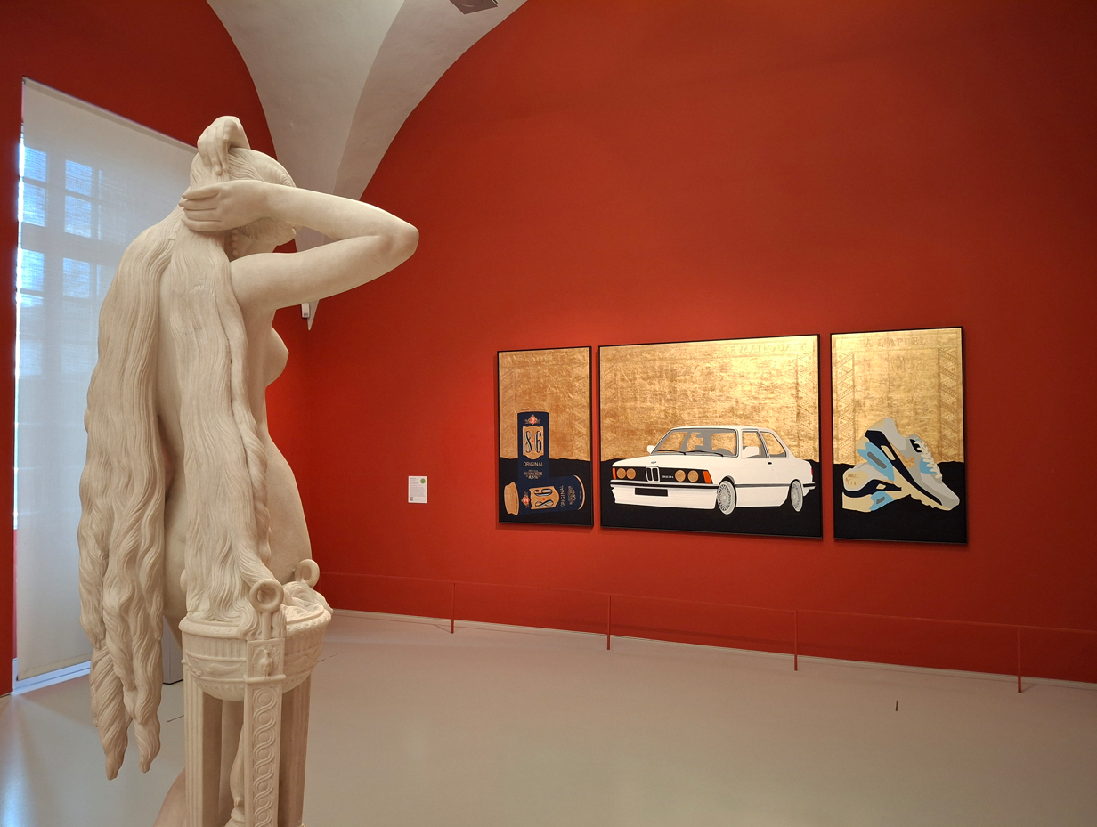 Une nymphe de marbre, face à un panneau de bois gravé représentant des baskets, une bmw et des canettes de bière