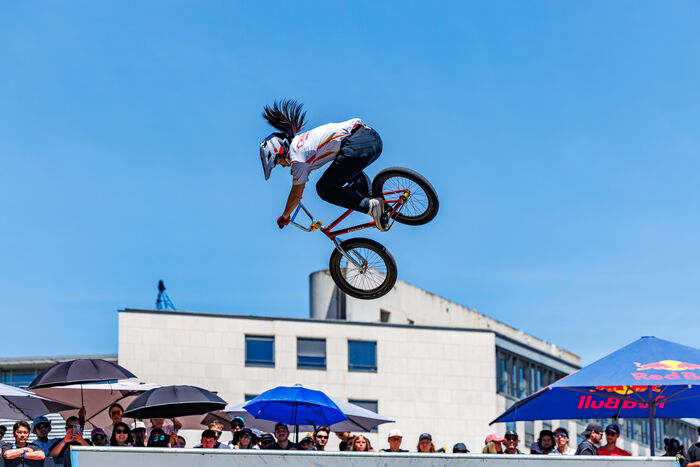 Bikeuse dans les airs avec son vélo, ciel, tribune, parasols publicitaires, batîment