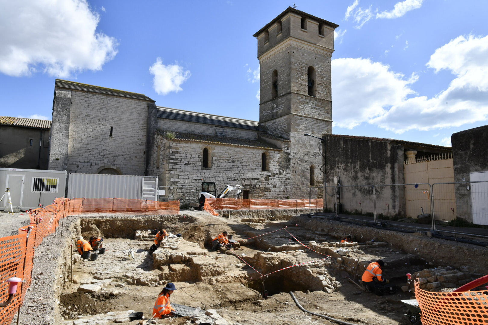 site de fouilles, archéologues, clôture de protection, église du XIe siècle en arrière-plan, ciel