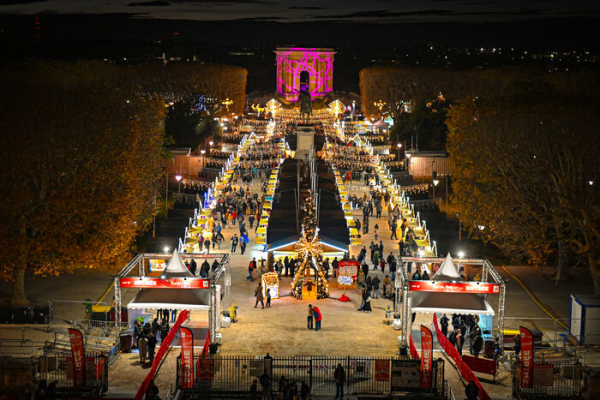 Vue surplombée du marché avec entrée, allées avec de nombreuses personnes, illuminations, chalets, château d'eau, végétation