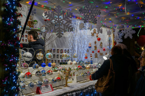 Commerçant de dos, chalet vendant des décorations de Noël, boules de Noël au premier plan, clients 
