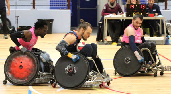 trois joueurs de rugby fauteuil en action sur le parquet, table de marque en arrière plan